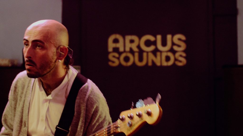 Bassist performing during the familiar action live session, in the background, the gold arcus sounds logo is visible on the purple music studio door, a london music studio
