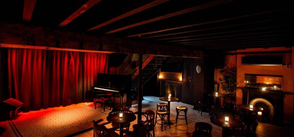 The downstairs of Servant's Jazz Quarters in East London, there is a stage with a piano and a fwe candlelit tables